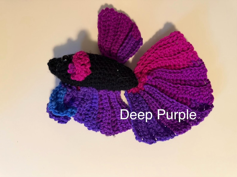 May include: A handmade crochet betta fish in shades of purple, magenta, and blue. The fish has a black head with a pink flower accent and the text "Deep Purple". This decorative item is a unique handmade gift.