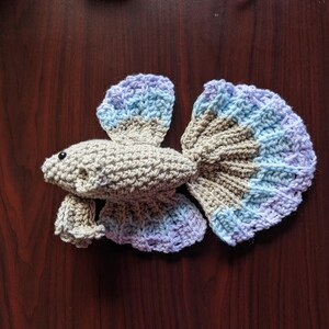 May include: A crocheted betta fish in shades of beige, blue, and purple. The fish has a detailed tail and fins, with a small black eye. The handmade fish is lying on a dark wood surface.