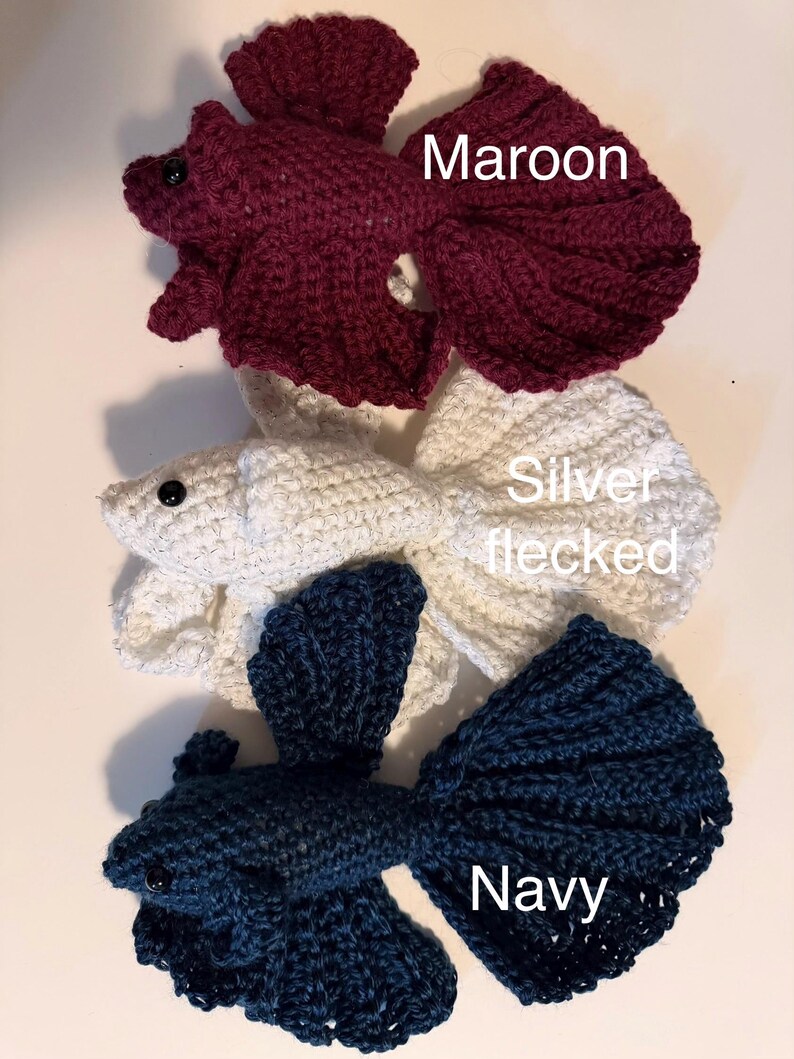 May include: Three crocheted betta fish in maroon, silver flecked, and navy blue. Each fish has a detailed tail and fins, with a small black eye. The fish are handmade and appear to be soft and plush.