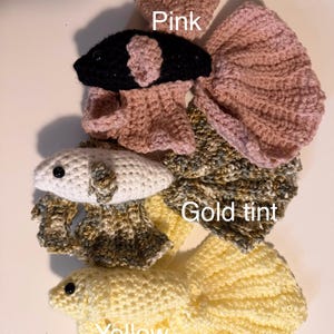 May include: Handmade crochet betta fish in pink, black, white, gold tint, and yellow. The image shows several fish with detailed fins and bodies. The word "Pink", "Gold tint", and "Yellow" are written on the image.