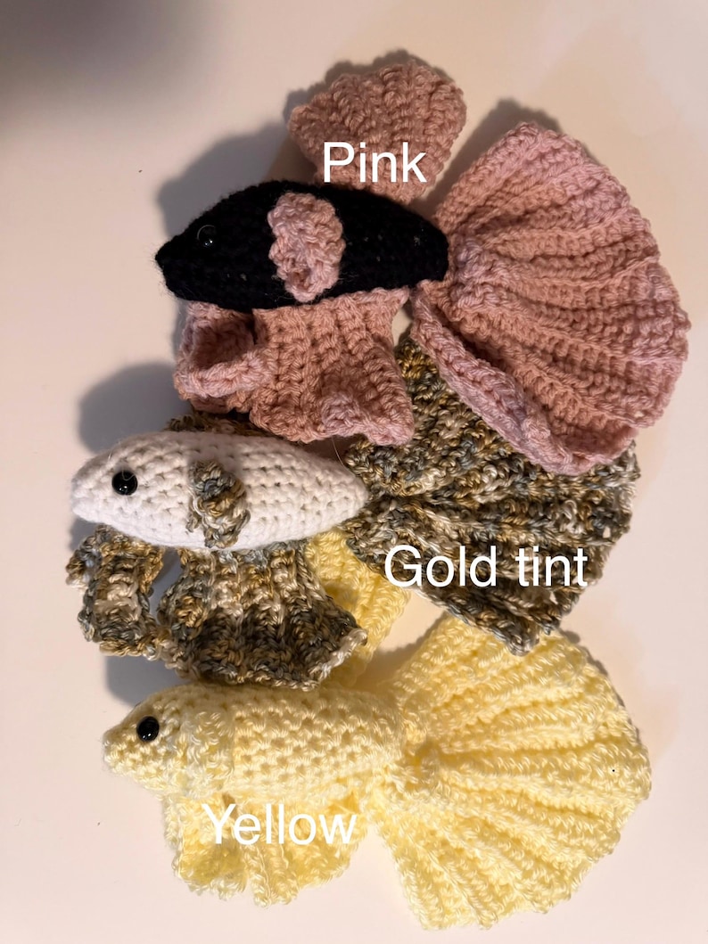 May include: Handmade crochet betta fish in pink, black, white, gold tint, and yellow. The image shows several fish with detailed fins and bodies. The word "Pink", "Gold tint", and "Yellow" are written on the image.