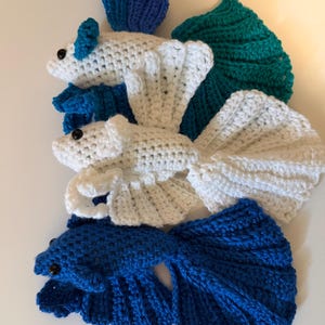 May include: Crocheted betta fish in shades of white, blue, and teal. The handmade fish have detailed fins and tails, with small black bead eyes. The fish are arranged on a white surface.