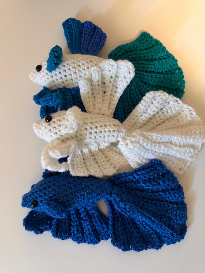 May include: Crocheted betta fish in shades of white, blue, and teal. The handmade fish have detailed fins and tails, with small black bead eyes. The fish are arranged on a white surface.