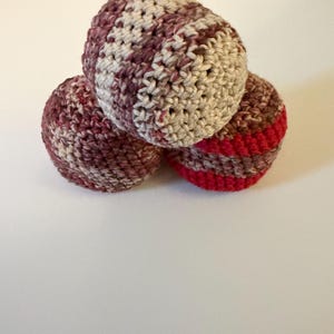 May include: Three handmade crochet balls in shades of burgundy, beige, and red are stacked. The balls have a textured appearance due to the crochet stitch. The balls are likely used for play or decoration.