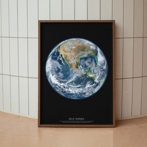 Puede incluir: Una impresión de arte mural rectangular enmarcada que presenta una imagen de alta resolución de la Tierra sobre un fondo negro sólido. El planeta muestra continentes, océanos y formaciones nubosas visibles. El texto BLUE MARBLE está impreso abajo.