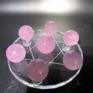 May include: Seven pink star rose quartz spheres arranged on a clear glass stand with a geometric design. The spheres are of various sizes and have a translucent appearance.