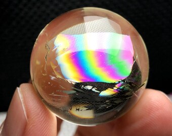 May include: A clear quartz crystal sphere with iridescent rainbow inclusions. The sphere is approximately 6.5 cm in diameter and is held in a hand.