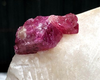 May include: A deep pink ruby crystal with a rough, faceted surface. The crystal is sitting on a white rock.