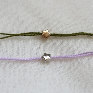 May include: Two thin cord bracelets, one in olive green with a gold-toned star charm and one in light lavender with a silver-toned star charm. Each bracelet features a single small star-shaped bead centred on a delicate, twisted string cord.