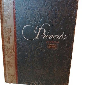 May include: A journal with a dark blue cover featuring an embossed floral pattern. The word "Proverbs" is written in elegant script, with "JOURNAL" below. The spine has a decorative design with a red accent.