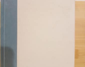 May include: A hardcover book with a light blue spine and the word "BECOMING" in gold lettering on the front cover. The cover is primarily off-white, with a clean and minimalist design. The book is set against a light wooden surface.