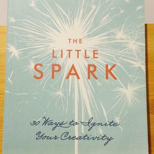 May include: A teal blue book with a white sparkler design on the cover. The title is "The Little Spark" and the subtitle is "30 Ways to Ignite Your Creativity". The author is Carrie Bloomston.