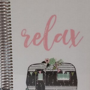 May include: A white notebook with a black and white illustration of a camper van with pink flowers and a banner. The word "relax" is written in pink cursive on the cover.