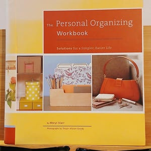 May include: A softcover book titled The Personal Organizing Workbook: Solutions for a Simpler, Easier Life by Meryl Starr. The cover features a yellow and orange color scheme with three inset photographs showing organized storage, office supplies, and a handbag.