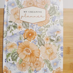 May include: A floral planner with a cover featuring orange and blue flowers, green leaves, and the text "MY CREATIVE planner" in a decorative font. The planner is on a wooden surface.