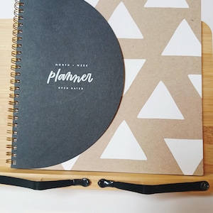 May include: A spiral-bound planner with a black cover and the words "Month + Week Planner Open Dated" in white. The other half of the planner has a tan background with white triangle patterns. A black elastic band lies below the planner.