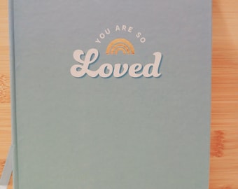 May include: A light blue self-care planner with a gold foil design that says "You are so Loved" with a rainbow graphic.