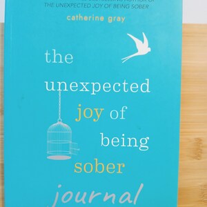 May include: A turquoise blue book cover with the title "the unexpected joy of being sober journal" in white text. The author's name, "catherine gray", is printed in white text above the title. A white bird is flying above the title and a birdcage with the door open is hanging below the title.