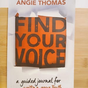 May include: A book cover with the title "FIND YOUR VOICE" in large, dark letters against an orange background. The author's name, Angie Thomas, is above the title. The cover also includes the text "a guided journal for writing your truth."