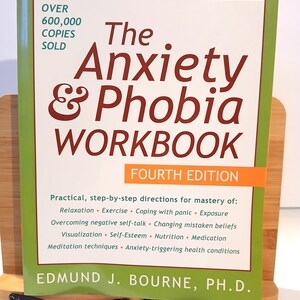 May include: A self-help workbook titled "The Anxiety & Phobia Workbook" with a green border and white background. The cover includes the text "Over 600,000 Copies Sold" and "Fourth Edition". The book is propped up on a wooden stand.