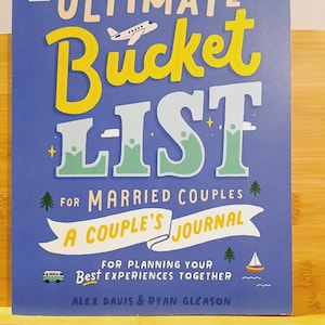 May include: A blue "Ultimate Bucket List" journal for married couples. The cover features the title in yellow and white, with illustrations of a plane, trees, and a sailboat. The text "A Couple's Journal" is also included.