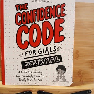 May include: A journal titled "The Confidence Code for Girls" with red and black text on a white background. The book is propped up on a bamboo book stand with black metal supports. The cover includes the authors' names and a tagline.