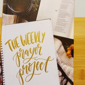 May include: A white journal with gold lettering that reads "The Weekly Prayer Project: A Challenge to Journal, Pray, Reflect". The journal is open to a blank page and is surrounded by a cup of coffee, a Bible, glasses, and a knitted blanket.