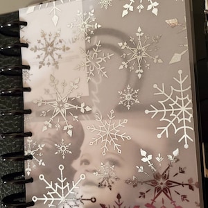 May include: A clear plastic sheet with a silver snowflake pattern. The sheet is designed to be used as a decorative cover for a planner or notebook.