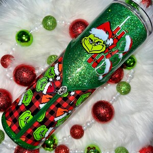 May include: A green glitter tumbler with a red and black plaid design featuring the Grinch. The tumbler has a green lid and a zipper detail. The Grinch is wearing a red Santa hat and the text "HO HO HO" is visible on the tumbler.
