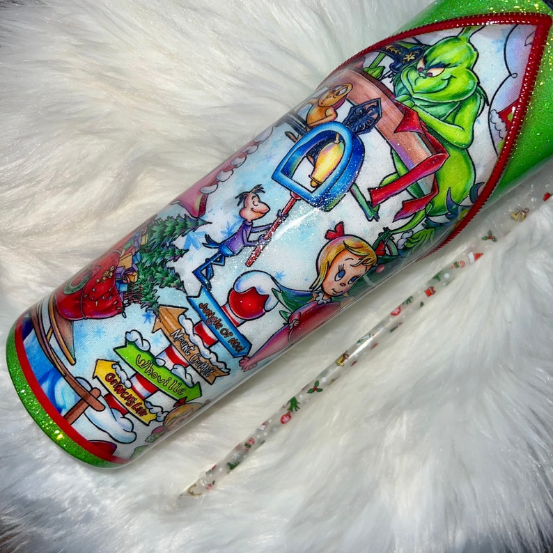 K&ouml;nnte beinhalten: Ein wei&szlig;er Becher mit einem gr&uuml;nen Glitzerrand und einem gr&uuml;nen und roten Glitzerstrohhalm. Der Becher zeigt eine farbenfrohe Illustration des Grinchs und Cindy Lou Who aus dem Film "Wie der Grinch Weihnachten gestohlen hat". Die Illustration enth&auml;lt Text, der "Whoville", "Nordpol", "Grinch" und "Weihnachtsmann" lautet.