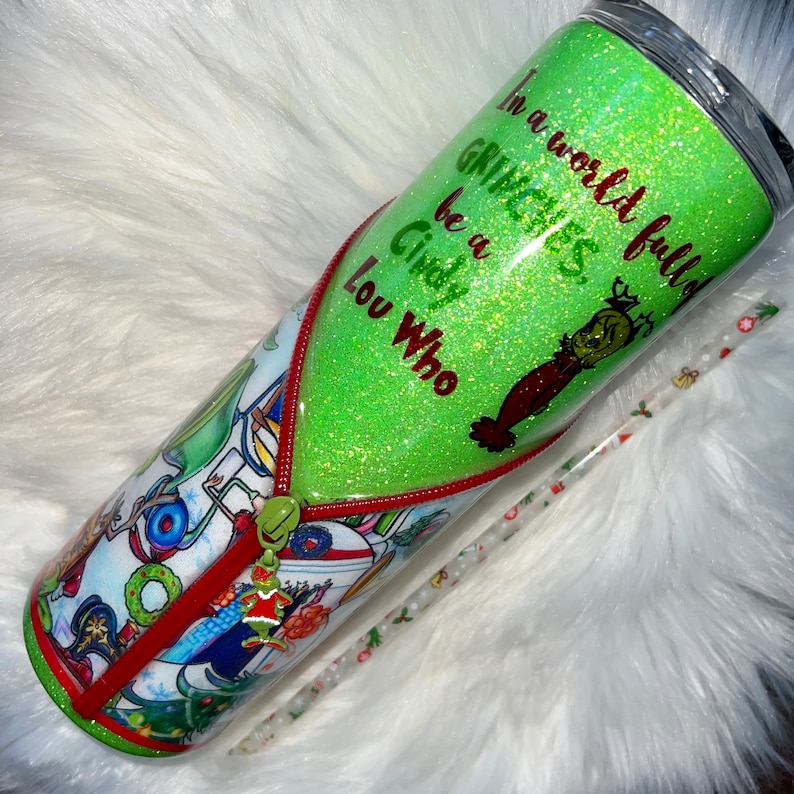 K&ouml;nnte beinhalten: Gr&uuml;ner Glitzerbecher mit rotem Rei&szlig;verschluss-Design und einer Grinch-Grafik. Der Becher tr&auml;gt den Text "In einer Welt voller Grinches, sei ein Cindy Lou Who".