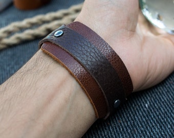 May include: A brown leather bracelet with a dark brown leather band on top. The bracelet has two silver metal snaps.