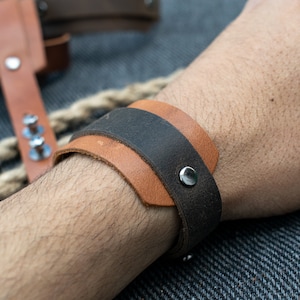 Puede incluir: Un brazalete de cuero marrón con una banda de cuero negro en la parte superior. El brazalete tiene una hebilla de metal plateado.