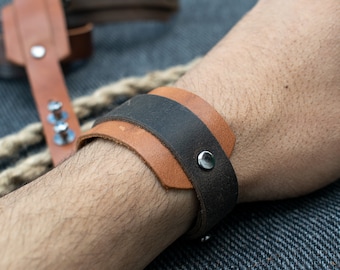 May include: A brown leather bracelet with a black leather band on top. The bracelet has a silver metal buckle.