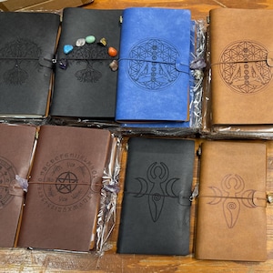 May include: A collection of rectangular journals with leather-like covers in black, brown, and blue. Each journal features a unique embossed design, including tree of life, geometric patterns, and celestial symbols, secured with a thin elastic band.