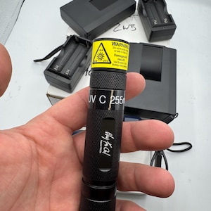 May include: A black cylindrical UV-C torch with a yellow warning label at the top, text reading UV C 255nm, and a logo with the text Ray to Col. The device includes two black battery chargers and a black rectangular case in the background.