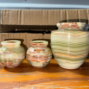 May include: Three decorative vases made of green, brown, and white striped onyx. The vases are different sizes and are arranged in a row on a wooden surface.
