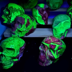 May include: A collection of skull-shaped objects featuring a marbled pattern of vibrant neon green and deep purple, glowing under ultraviolet light. A yellow ruler is visible in the lower right corner, showing a measurement of approximately 2 inches.