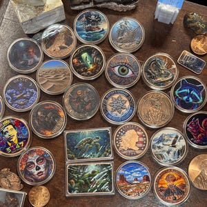 May include: A collection of circular and rectangular metal tokens featuring diverse colorful illustrations including a pirate ship, a desert landscape, a UFO, a dragon, a raccoon, a squirrel, a skull, and a portrait of a person with the text 5247.