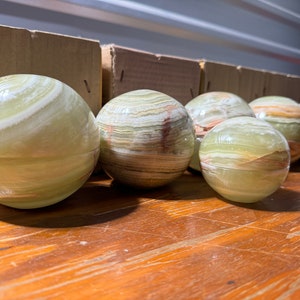 May include: Six green and white striped marble spheres, with some spheres having brown stripes, are arranged in a row on a wooden surface.