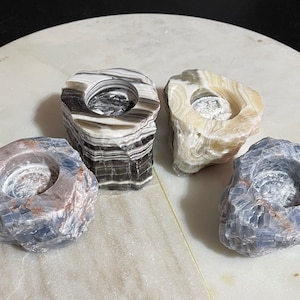 May include: Four natural stone candle holders in different colors and patterns. The holders are made of onyx, selenite, and blue calcite. They are all roughly the same size and shape, with a round opening in the center for a candle.