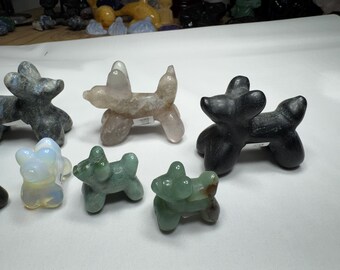 May include: A collection of seven small, hand-carved stone sculptures of dogs. The sculptures are in various colours, including black, grey, white, green, and pink. They are all in the shape of a dog with its legs stretched out.