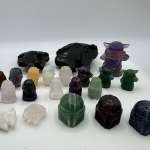May include: A collection of hand-carved crystal figurines of Star Wars characters, including Yoda, Grogu, and Mandalorian helmets. The figurines are made from various types of crystals, including amethyst, clear quartz, and green aventurine.