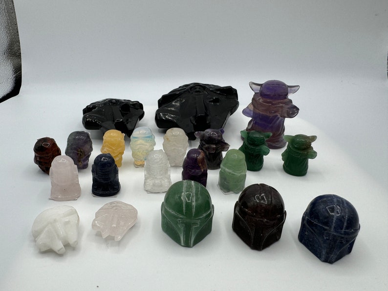 May include: A collection of hand-carved crystal figurines of Star Wars characters, including Yoda, Grogu, and Mandalorian helmets. The figurines are made from various types of crystals, including amethyst, clear quartz, and green aventurine.