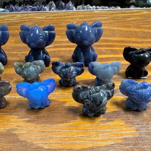 May include: A collection of small, carved stone figurines in various colors, including blue, gray, and black. The figurines are in the shape of a stylized animal with large ears. They are arranged on a wooden surface.