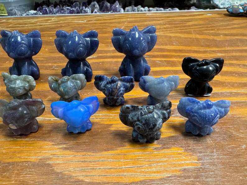 May include: A collection of small, carved stone figurines in various colors, including blue, gray, and black. The figurines are in the shape of a stylized animal with large ears. They are arranged on a wooden surface.