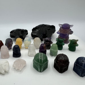 May include: A collection of hand-carved crystal figurines of Star Wars characters, including Yoda, Grogu, and Mandalorian helmets. The figurines are made from various types of crystals, including amethyst, clear quartz, and green aventurine.