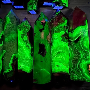 May include: A collection of glowing green crystal obelisks with detailed patterns and dark brown and red accents. The crystals are lit up, highlighting their unique internal structures. The crystals are displayed on a dark surface.