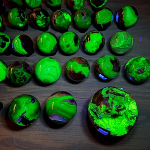 May include: A collection of spherical stones arranged in rows, each displaying a vibrant, glowing neon green pattern against dark brown or black areas. The surfaces are smooth and polished, reflecting light to highlight the intense fluorescent green color.