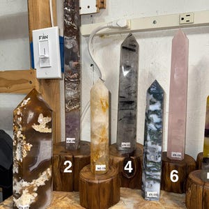 Può includere: Una collezione di sei obelischi di cristallo lucido di vari colori e motivi, esposti su supporti di legno. I cristalli includono tonalità marroni, viola, trasparenti, nere, verdi e rosa. Ogni obelisco ha una parte superiore a punta.